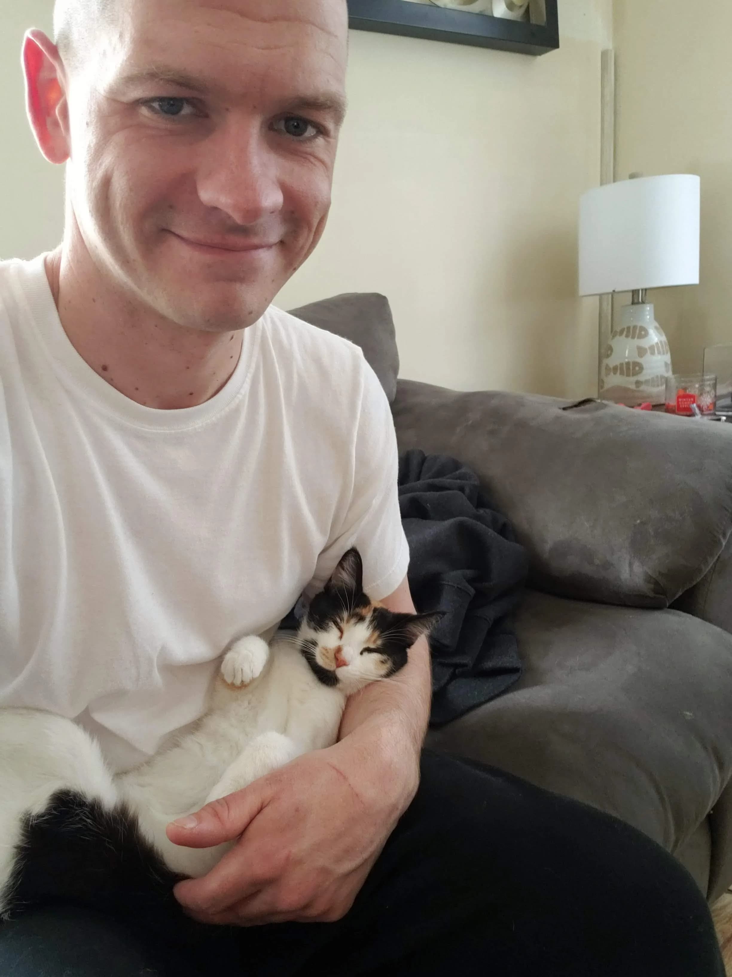 Sean in a white vest holding Bonnie as a kitten against his chest, one hand cupped under her, both looking toward the camera