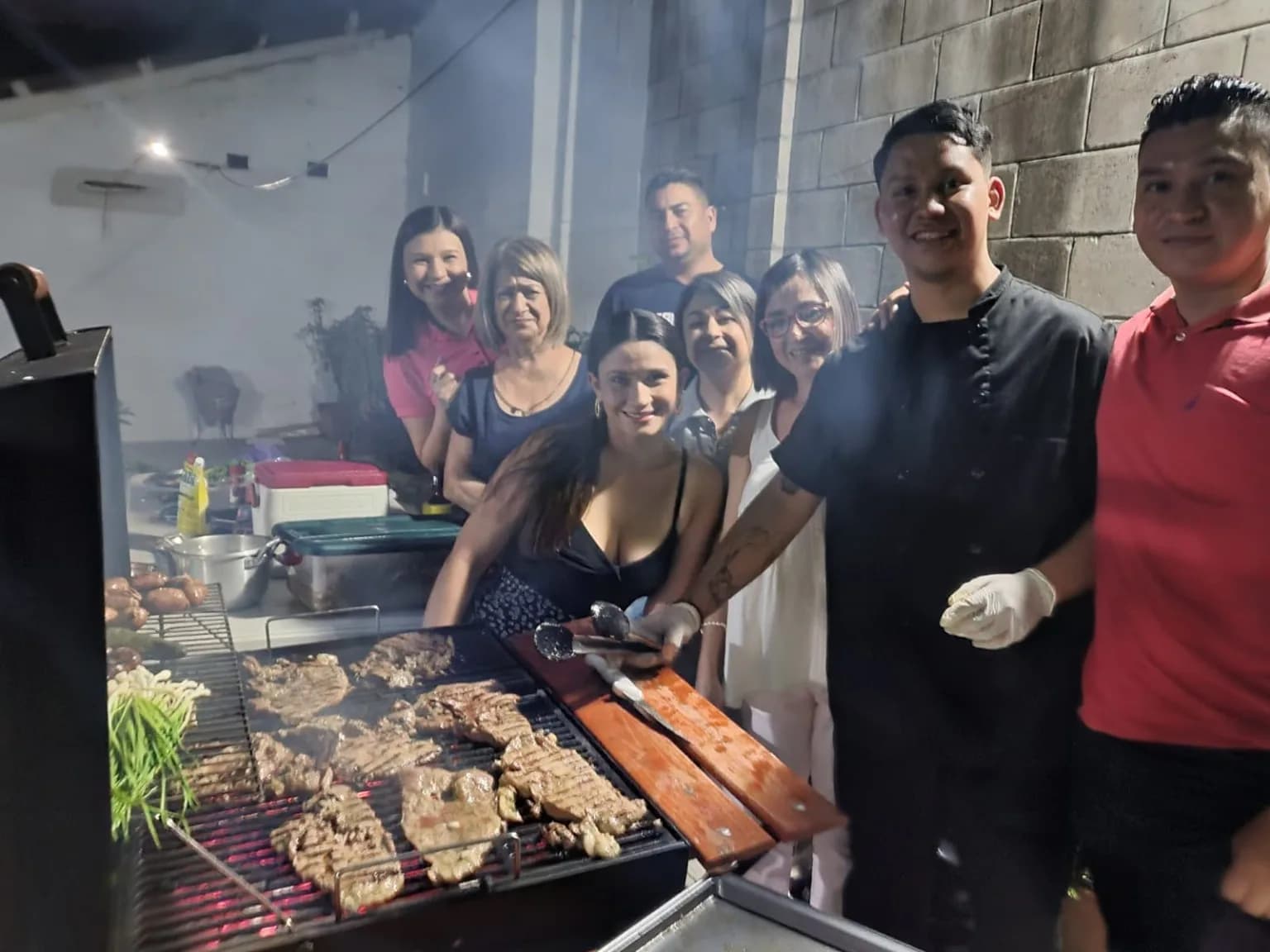 The family gathered around a full grill in El Salvador, meat and green onions cooking, everyone turning to face the camera