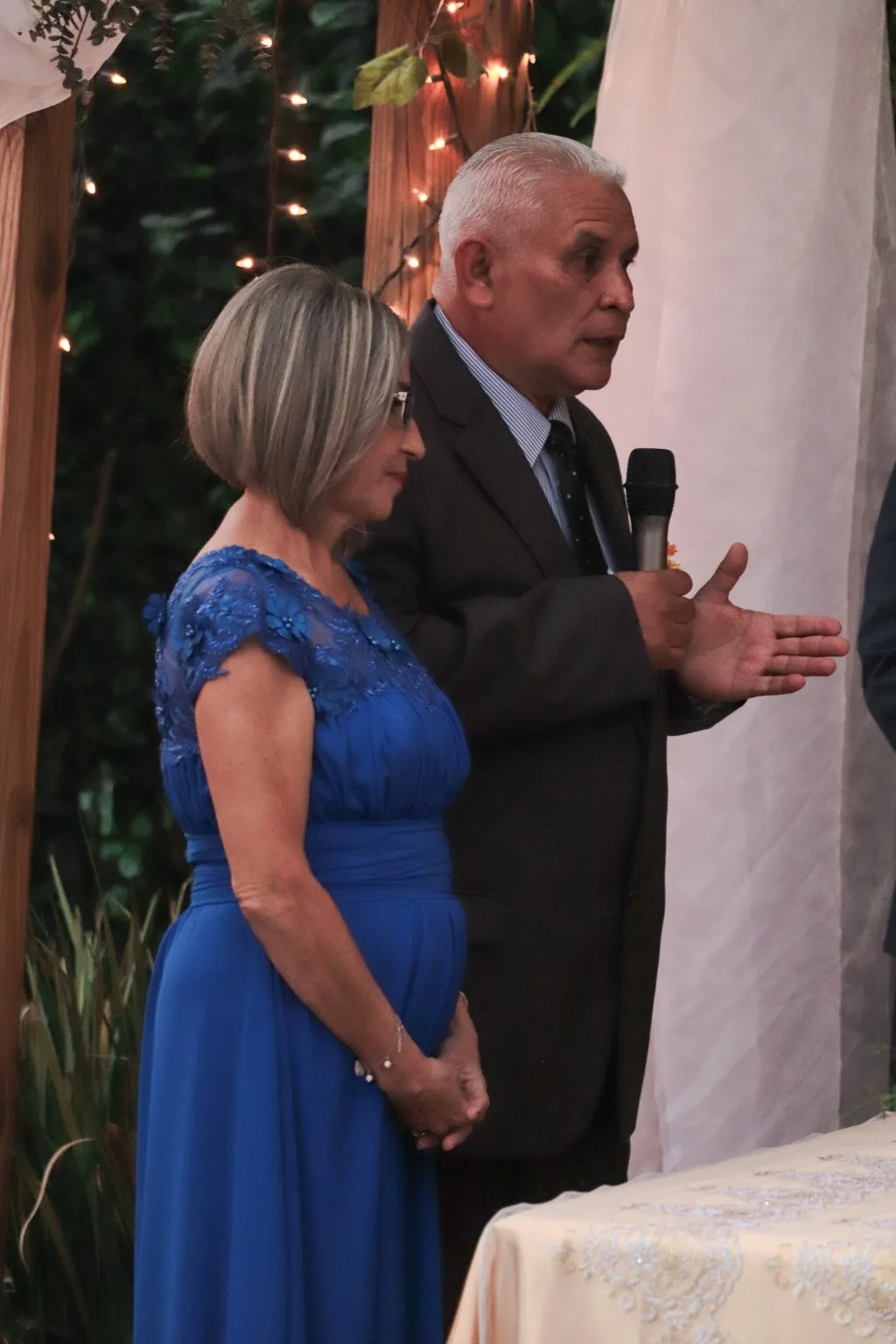 Florencia's grandparents at the wedding, her abuelo speaking into a microphone while her abuela listens beside him
