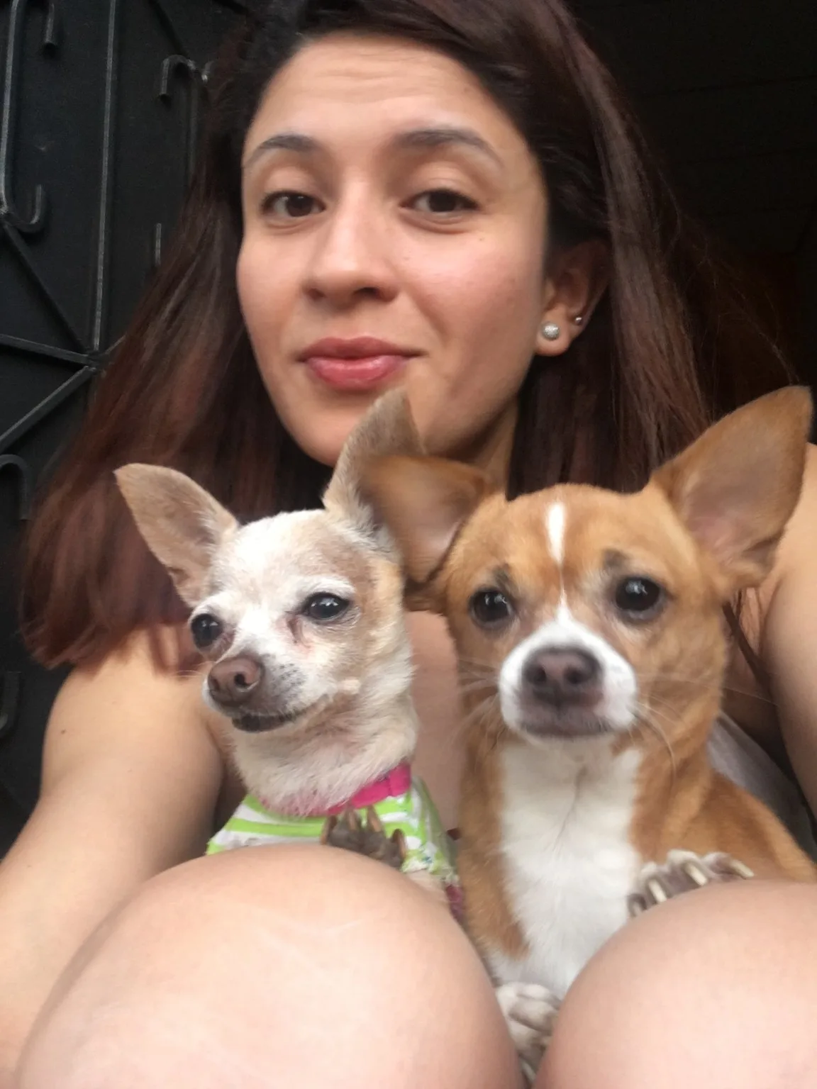 Florencia with Chanel, a tiny cream Chihuahua in a striped outfit, and Bruno, a brown-and-white Chihuahua, sitting together on her lap in front of a dark iron door